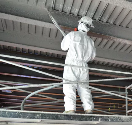 Worker in a white protective suit installing fireproofing material to a white ceiling.