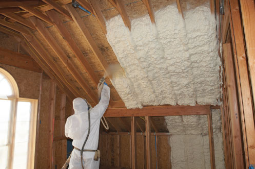 Worker in a white protective suit installing spray foam onto interior walls and slanted ceiling.