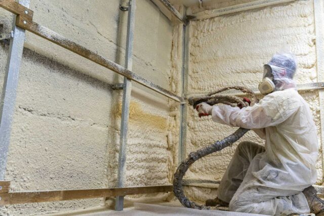 Worker installing spray foam insulation on walls in a metal building.