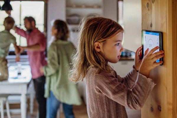 Little girl adjusting a smart thermostat on home's wall, with family in the background.