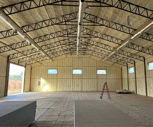 Interior of a large metal building showing professional installation of closed-cell spray foam insulation on the walls and ceiling. The space features exposed metal trusses, HVAC ductwork, and an open bay door.