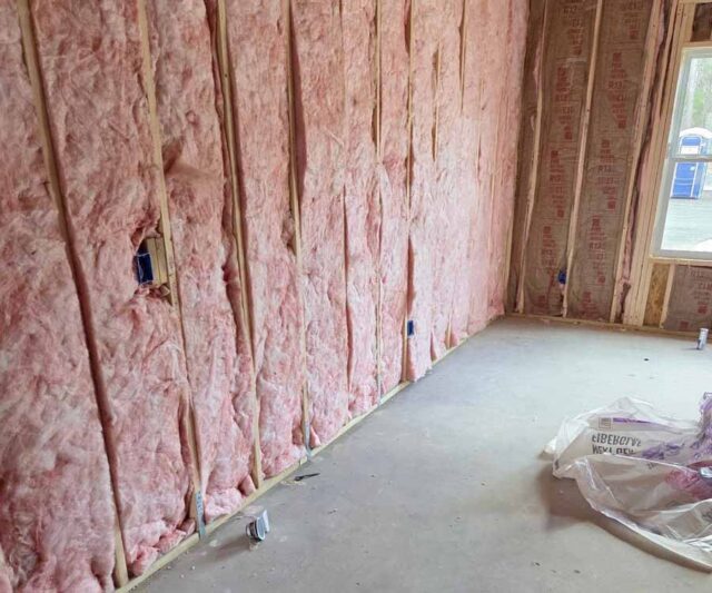 An interior view of a room under construction with pink fiberglass batt insulation installed between vertical wooden wall studs.