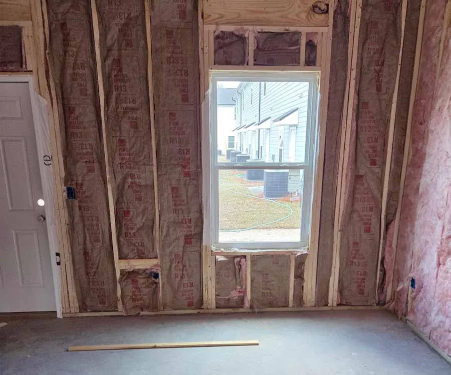 An interior wall of a room under construction, showing pink fiberglass batt insulation with brown kraft paper facing installed between wooden studs.