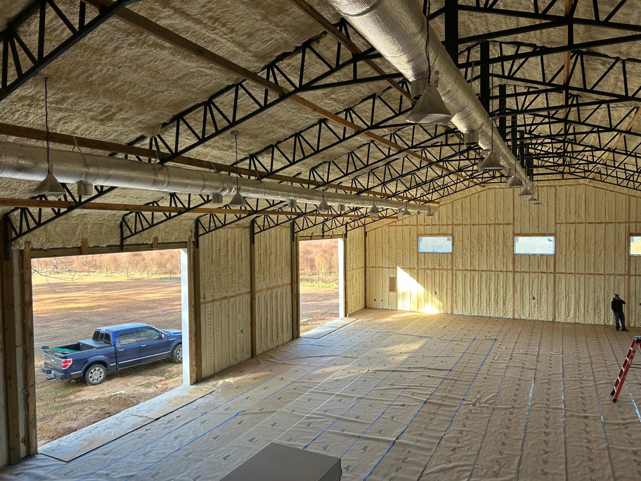 Professional spray foam insulation installation in a large metal shop building with open bay doors and a blue truck parked outside.