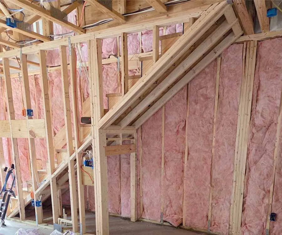 An interior view of a house under construction showing the wooden framing of a staircase. Pink fiberglass batt insulation is installed between the vertical wall studs and within the cavities under the stairs.
